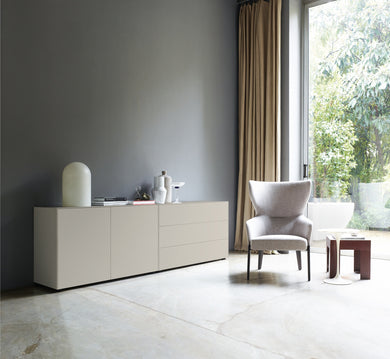Seidengraue Sideboardkombination in einer stilvollen Einrichtung mit grauer Wand, Vorhängen und Sessel neben großem Fenster, mit minimalistischer Dekoration.